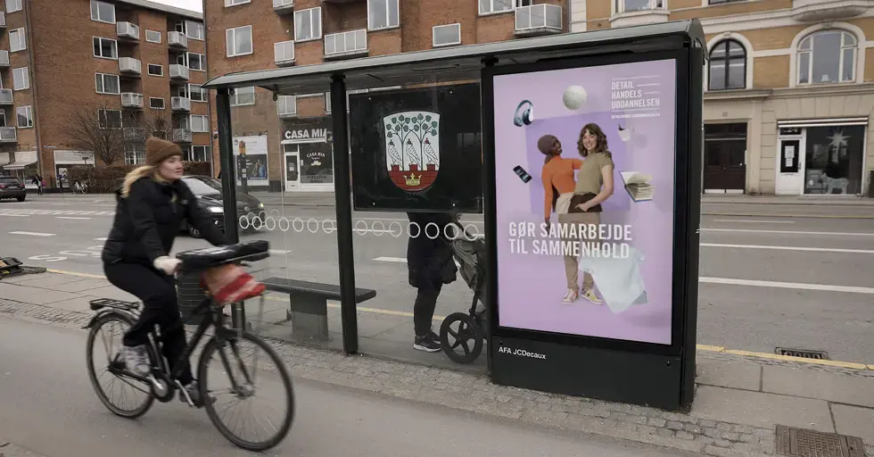Busskur med plakat, hvor to unge samarbejder. Cyklist og bygninger i baggrunden.