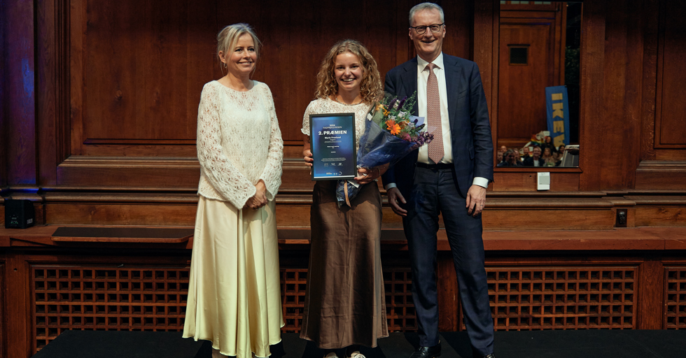 Ung kvinde modtager pris og blomster flankeret af to ældre personer på scene i auditorium