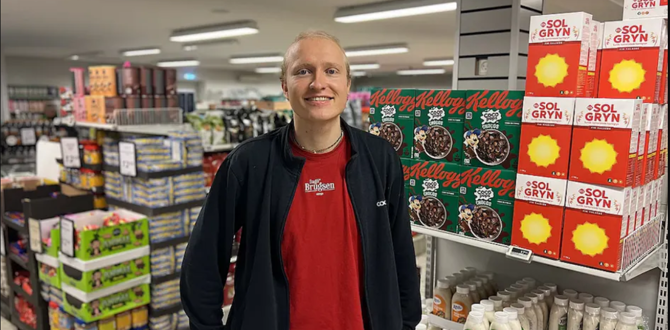Medarbejder i rød t-shirt og mørk jakke står smilende i en dagligvarebutik omgivet af hylder med varer