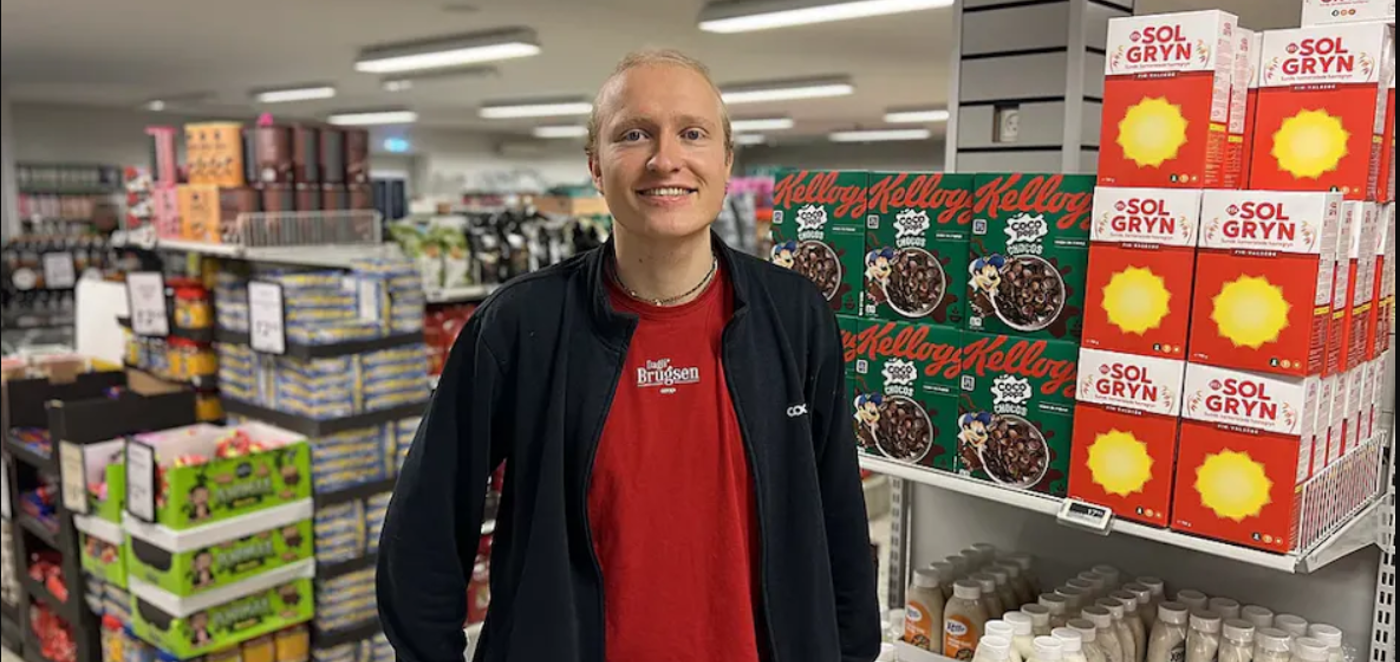 Medarbejder i rød t-shirt og mørk jakke står smilende i en dagligvarebutik omgivet af hylder med varer