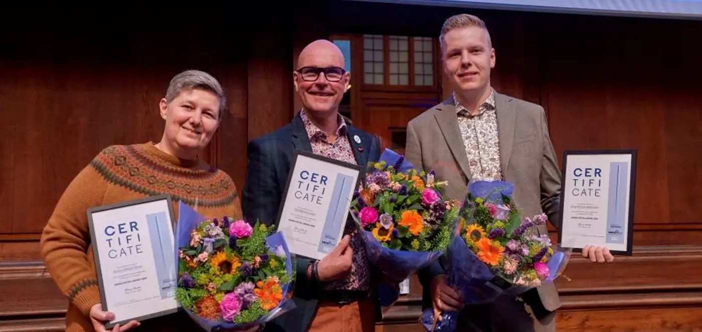 Tre personer står på en scene med certifikater og farverige blomsterbuketter