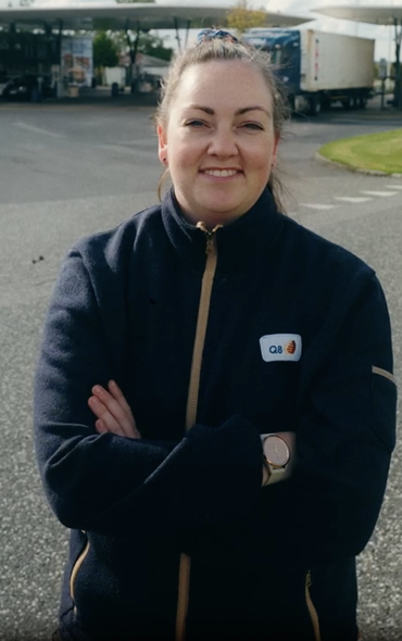 Kvinde i mørkeblå fleece med Q8-logo smiler til kameraet på en parkeringsplads