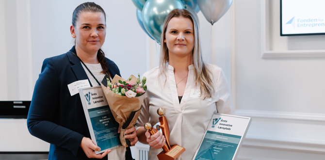 To kvinder holder diplomer, buket og statuette ved prisoverrækkelse med balloner i baggrunden