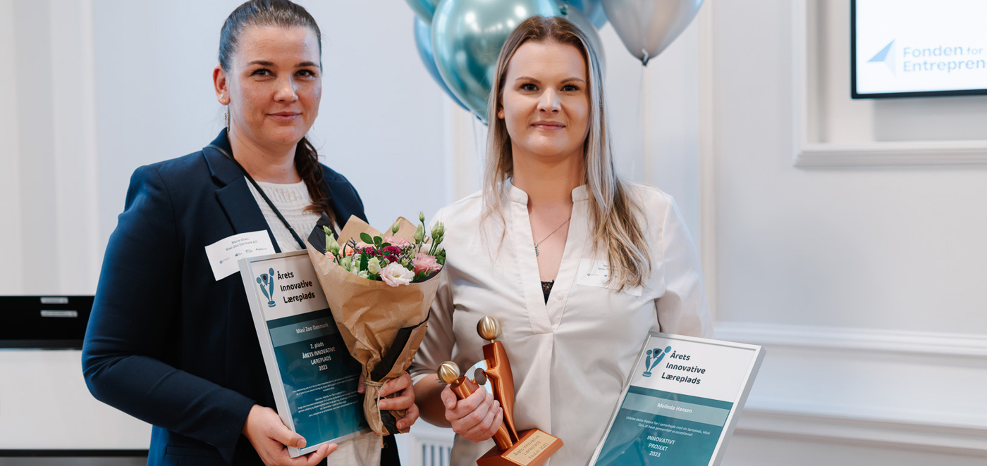 To kvinder holder diplomer, buket og statuette ved prisoverrækkelse med balloner i baggrunden