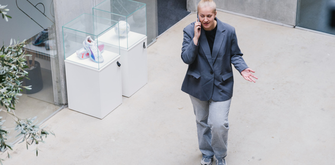Ung kvinde i jakkesæt taler i telefon mens hun går gennem moderne bygning med udstillingsvitrine