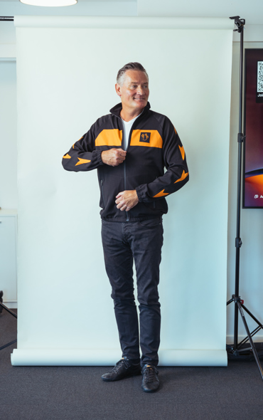 Smilende mand i sort og orange sports-træningsjakke foran hvid baggrund med fotoudstyr