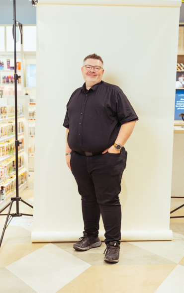 Mand i sort skjorte og briller står foran hvid baggrund med fotoudstyr og hylde med produkter