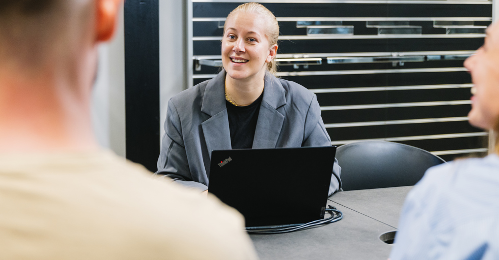 Ung kvinde i grå blazer sidder ved møde med laptop og smiler til kollegaer