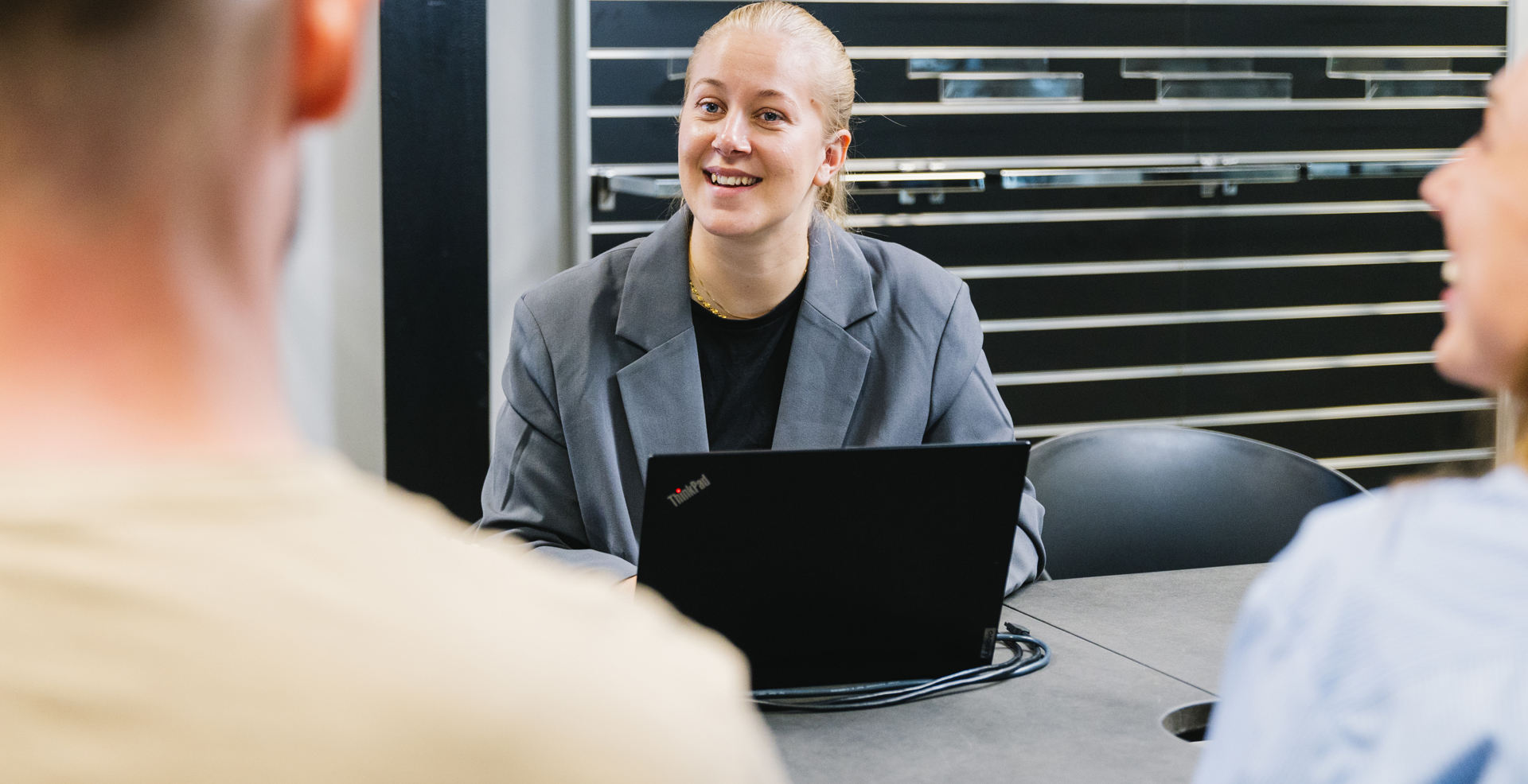 Ung kvinde i grå blazer sidder ved møde med laptop og smiler til kollegaer