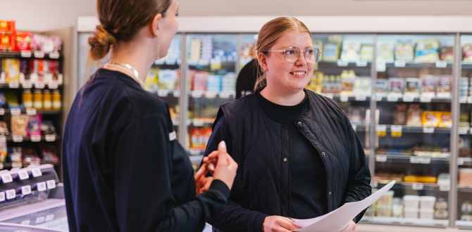 To unge kvinder i sort arbejdstøj taler sammen i en dagligvarebutik med kølediske i baggrunden