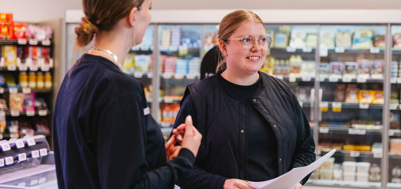 To unge kvinder i sort arbejdstøj taler sammen i en dagligvarebutik med kølediske i baggrunden