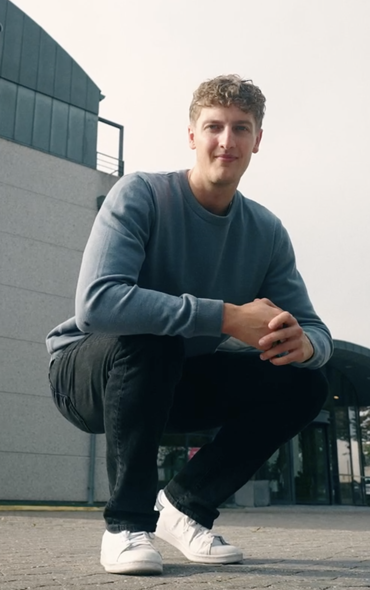 Ung mand i grå sweatshirt sidder på hug foran moderne skolebygning