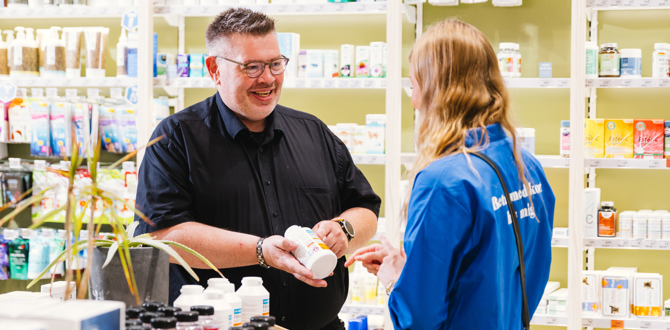 Medarbejder i sort skjorte vejleder elev i blå trøje om produkter i butik med hylder